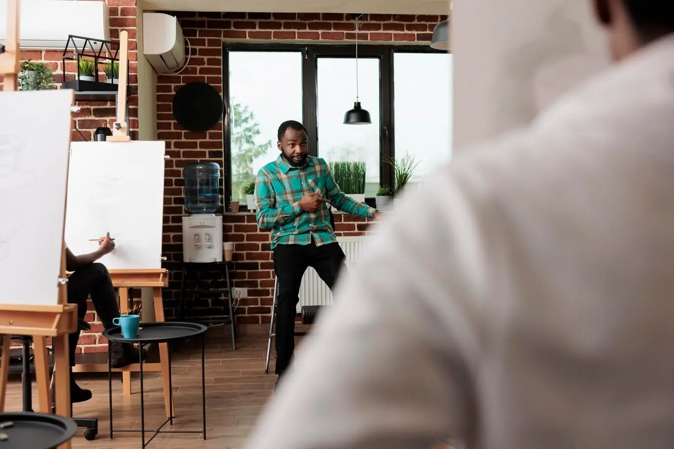 En un estudio creativo, un hombre joven afroamericano enseña arte; el instructor masculino demuestra técnicas de boceto a estudiantes sentados frente a caballetes durante un taller grupal de dibujo.